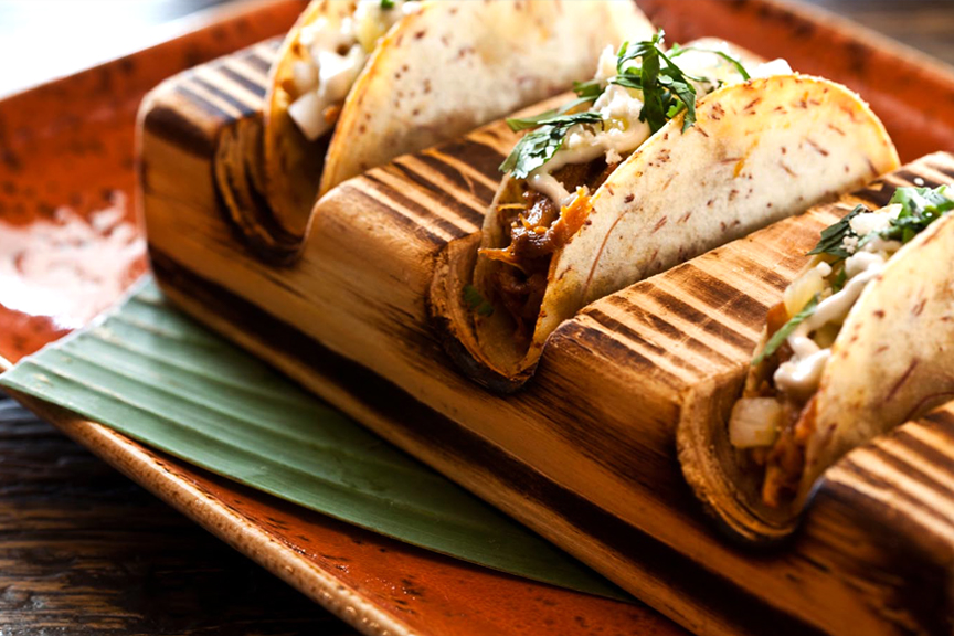 Focus on three gourmet Churrascos tacos, in a wooden taco holder, on a rust-colored clay dish.