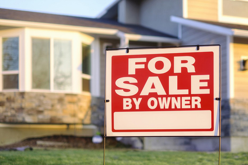 A red, in focus, “For Sale By Owner” sign standing in front of an out of focus single-family home.
