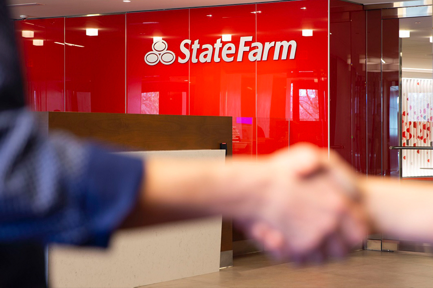 Office interior. Focus on a State Farm logo fashioned to a red, gloss wall. Diminishing focus on two hands shaking in the foreground.
