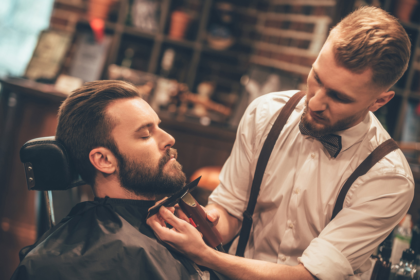 A man having his beard trimmed by a young barber wearing a white button down, suspenders, and a bow tie.