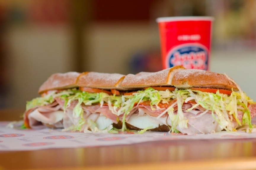 A Jersey Mike’s turkey and ham sub sandwich in the foreground. A red fountain drink featuring the company logo in the background.