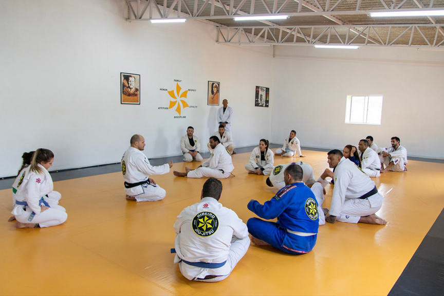 Six Blades Jiu Jitsu, bright, interior. Men and women sitting on the floor, listening to an instructor.