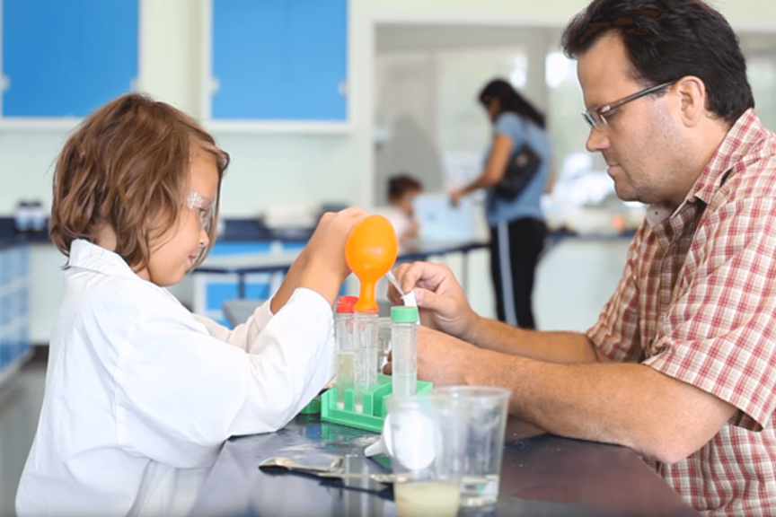 Father and son conducting an experiment with beakers and balloons inside a brightly lit room. Young boy wears a lab coat and safety glasses.